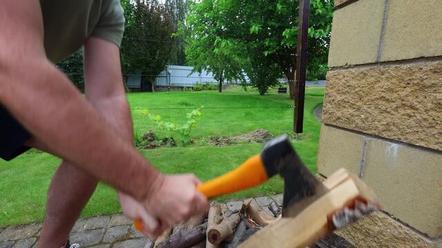 A man splits firewood with an orange-handled axe on a log in a garden, preparing fuel for grilling or outdoor cooking.