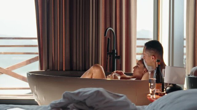 Romantic couple enjoying a hot bubble bath together while clinking champagne glasses in a rustic wooden cabin. Morning light pours in through the window, creating an intimate and serene atmosphere