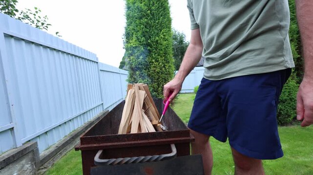 A man lights kindling in a rusty metal firebox outdoors, preparing a fire for grilling or heating in a backyard setting.