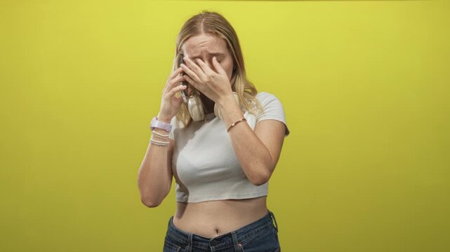Woman holding phone with hand covering mouth, wearing headphones, crop top and jeans in studio with yellow backdrop; distress call.