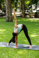 Young Asian woman practicing yoga in triangle pose on mat at green park. Healthy female doing stretching exercise outdoors for flexibility.