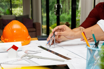 Close up of engineer hand using compass divider on construction floor plan. Architectural design concept with orange safety helmet and house model on desk.