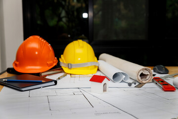 Construction workspace with orange and yellow safety helmets, rolled blueprints, and house model. Engineering desk setup with spirit level and notebook.