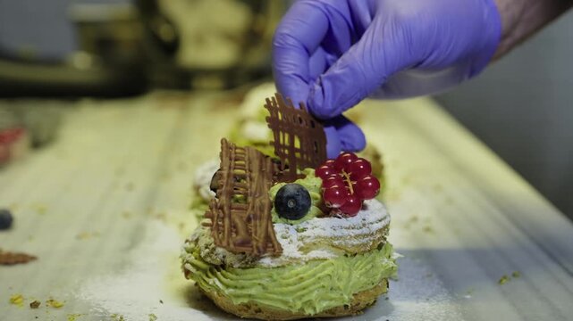 Pastry chef meticulously decorating a pistachio paris-brest with chocolate, blueberries, and red currants in a professional bakery kitchen for a gourmet dessert