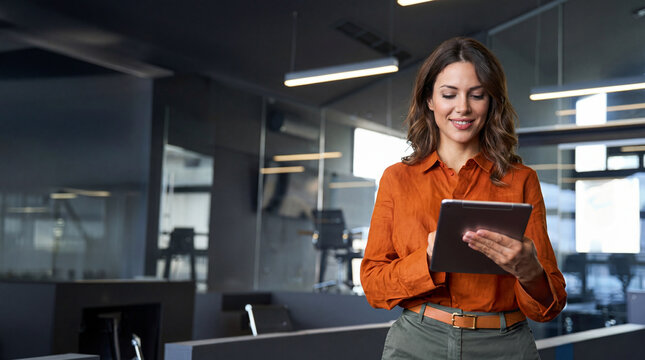 Mature adult professional business woman looking at online trade app. European businesswoman CEO holding digital tablet using fintech tab application standing at workplace in office.