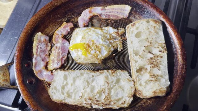 Closeup overhead shot of an egg, three rashers of bacon and two halves of panini bread, cooking in a frying pan.