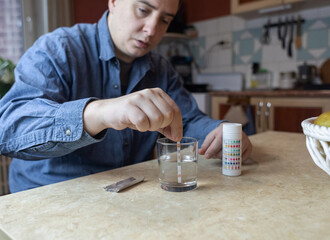 A man in the kitchen tests tap water quality with a test strip. Home testing for drinking water safety, pH, chlorine, and chemical contaminants. © Siniehina