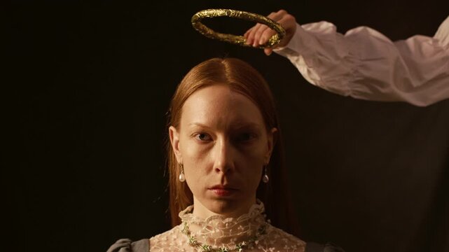 Portrait of young red haired woman wearing pearl earrings looking at camera while unseen person holding golden halo crown above queens head during conceptual shoot in studio against black background