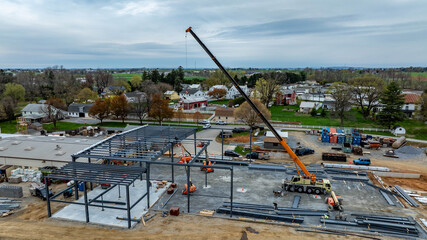 A crane is lifting materials at a construction site. Workers are present, and houses are visible in the background. The sky is cloudy, and the area is developing.