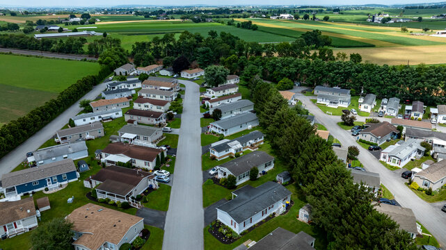 A community of Mobile, Manufactured houses arranged along winding streets. Green fields and farmland stretch in the background under a cloudy sky. It is a typical day in summer.