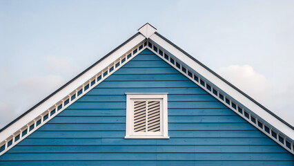 Obraz premium Blue wooden gable with white ventilation grille under pale sky, horizontal siding and roofline that emphasizes home airflow. 