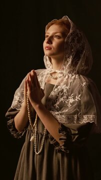Vertical shot of young red haired woman wearing white lace mantilla praying with rosary in hands in studio with warm low key lighting against back background, slow motion