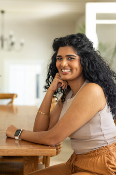 Indian woman sitting at wooden dining table leaning on hand, smiling while wearing smartwatch