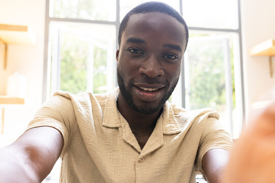 African American man in beige shirt holding phone, reaching toward camera at home by window shelves