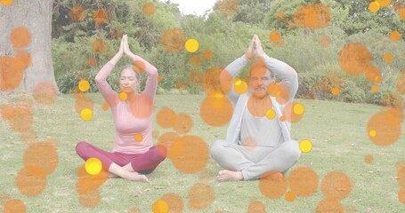 Meditating couple holding prayer pose on grass in park, wearing activewear with bokeh overlays