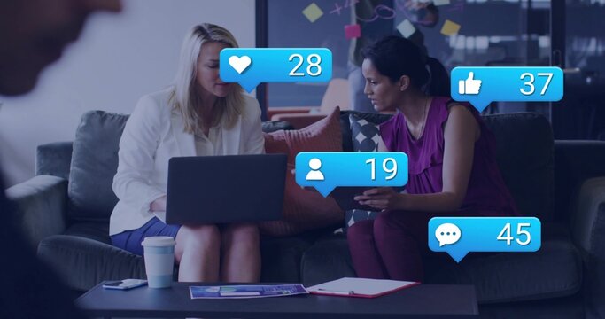 Reviewing laptop, tablet, two women in office lounge in white blazer and magenta top, social icons