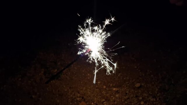 Handheld sparkler firework burning in the dark at night, glowing golden sparks from firecracker in pitch black background, cinematic light display for celebration and festive holiday atmosphere, close