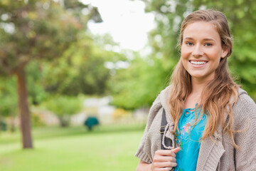 Fototapeta premium Female standing in park holding bag strap, wearing blue embroidered top, gray cardigan, copy space