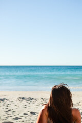 woman sitting on sandy shoreline facing calm turquoise ocean under clear blue sky