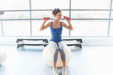 Fototapeta premium Woman sitting on white exercise ball in bright gym holding red dumbbells, wearing blue tank top