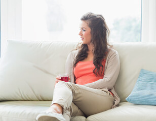 Fototapeta premium Woman sitting on sofa by window wearing coral top, beige cardigan holding glass with red drink