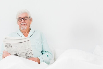 Senior man sitting in bed reading broadsheet newspaper wearing mint pajamas, glasses, copy space