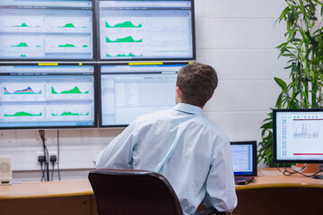 Curved wooden workstation displaying green charts on six wall screens, with potted plant near desk