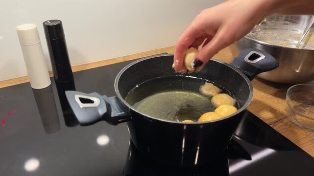 Adding Multiple Dough Balls to Deep Frying Pot. Hand quickly drops several rounds of dough into pot of bubbling oil. Induction stove and raw dough bowl are visible in background.