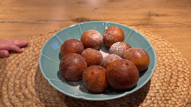 Close-up of Powdered Sugar Falling on Golden Pastries. Powdered sugar is dusted over bowl of warm golden-brown dough balls on woven mat. Fine white powder coats crispy exterior of homemade treats.