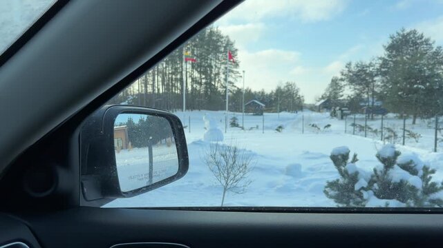 Looking Out Car Window at Snowy Winter Property. View through car window shows snow-covered yard with fence and modern buildings. Corgi dog stands alert in deep snow near gated driveway.