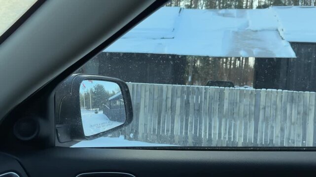 View from Car Driving Down Snowy Forest Lane. Camera captures roadside view from moving car passing wooden fence and dense pine forest blanketed in snow. Modern houses visible behind tree line.