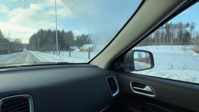 Car Driving on Paved Road Through Snow-Covered Area. Looking out from passenger side of car driving past long brown fence and snow-laden fields. Distant houses and forests complete serene landscape.