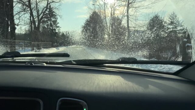 Windshield Wipers Clearing View on Sunny Winter Drive. Driver's perspective of traveling down snowy road while wipers clear moisture. Bright sun shines ahead illuminating wet asphalt and landscape.