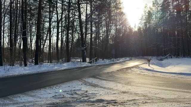 Wet Asphalt Road Curving Through Winter Forest. Quiet wet asphalt road curves through snow-covered forest. Low winter sun shines brightly through bare trees creating strong glare and reflections.