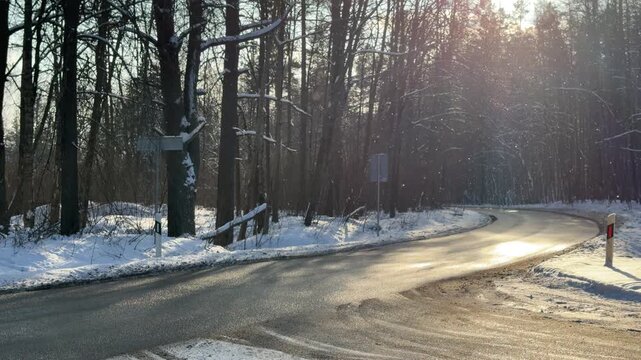 Cars Driving on Wet Curving Road in Winter Forest. Two cars drive around bend on wet asphalt road through snowy forest. Sun is low casting bright backlight and reflecting off road surface.