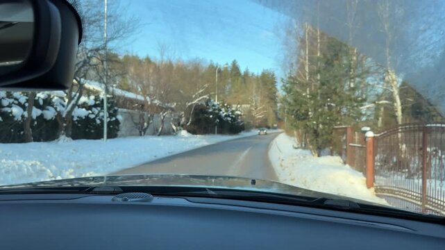 POV Driving on Winding Road in Snowy Village. Point-of-view shot from car driving along winding paved road through rural residential area. Snow banks line street with pine trees and houses visible.