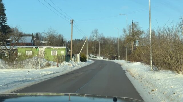 Driving Through Quiet Snowy Village on Sunny Day. POV from car traveling down straight road in small village. Features traditional houses and bare trees set against bright blue winter sky and snow.