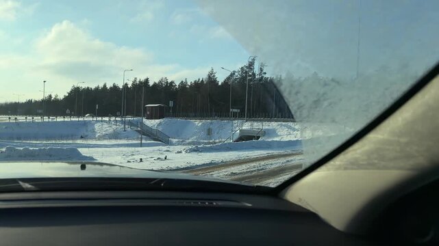 Approaching Roundabout on Snowy Winter Road. POV driving shot approaching traffic circle in rural area. Road signs and snow-covered fields visible through windshield catching low winter sunlight.