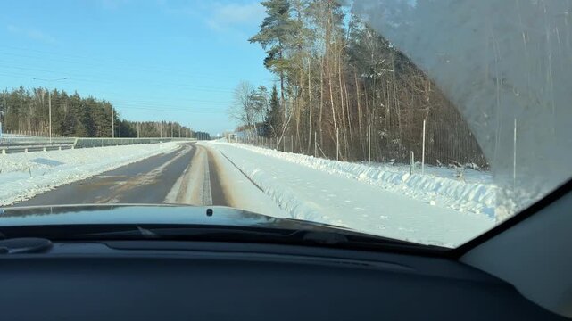Driving on Slushy Forest Road in Winter. POV shot from car traveling on two-lane road with slushy tracks in center. Road is surrounded by snow-covered forest under clear blue sky.