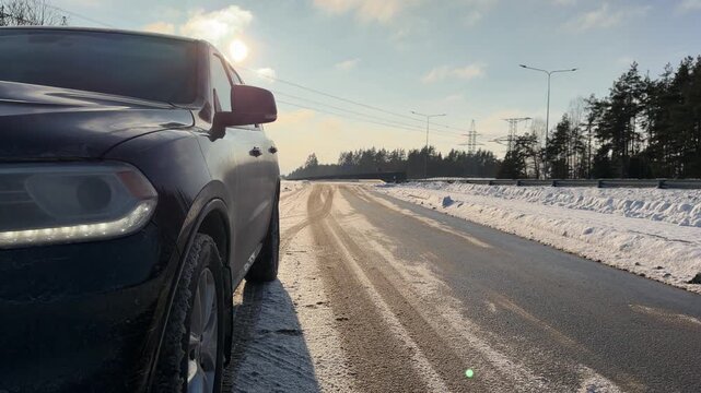 Sunlit Winter Sky and Parked SUV on Highway. Panning down from bright cloudy winter sky to black SUV parked on side of wet highway. Sun creates silhouette effect and lens flare over snowy landscape.
