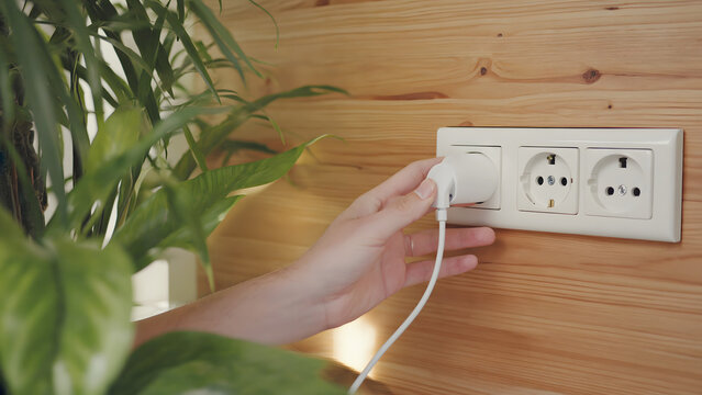 Hand plugging white charger into wall socket on wooden panel.