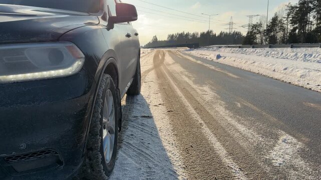 Front View of Car on Snowy Road Shoulder. Wide shot showing front of black SUV parked on snowy shoulder of highway. Sun creates backlight effect illuminating wet road and winter landscape.
