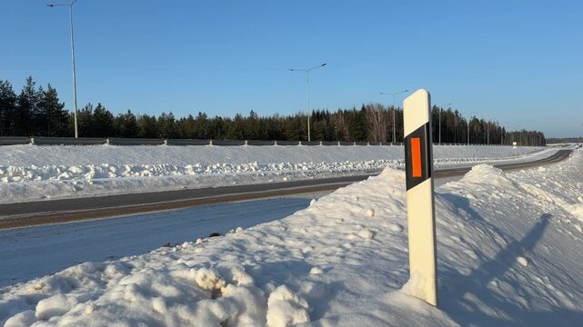 Road Marker Post and Traffic on Winter Highway. Static shot focusing on road reflector post on snowy embankment. Car drives past on highway in background highlighting winter travel conditions.