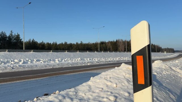 Roadside Reflector Post on Snowy Winter Highway. White road marker post with orange reflector stands on snow bank next to cleared highway. Cars pass by in background under clear blue sky.