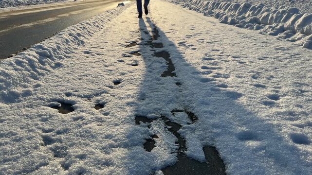 Walking on Snowy Sidewalk with Long Shadows. Low-angle shot of legs walking away on snow-covered paved path. Low winter sun casts long dramatic shadows of walker on sparkling white surface.