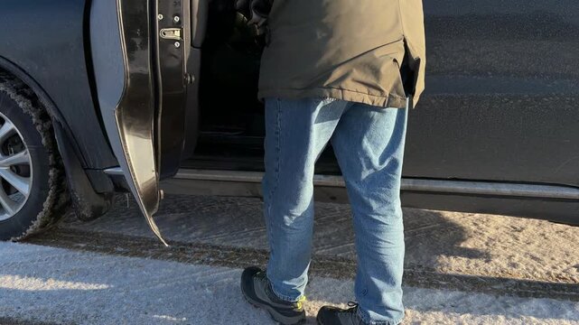 Driver Entering Dirty Car on Snowy Winter Day. Person wearing jeans and winter boots opens door of dirty black car and gets into driver's seat. Vehicle is parked on snowy roadside.