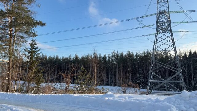 Winter Road with High Voltage Power Lines. Snow-covered rural road runs past large high-voltage electricity pylon. Background features pine forest and blue sky with soft clouds on sunny winter day.