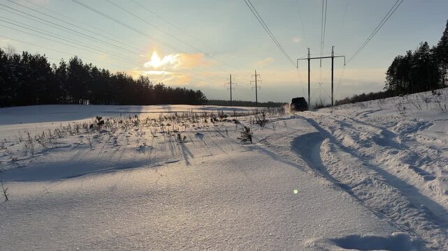 SUV Driving Off-Road in Snowy Field at Sunset. Black SUV drives through deep snow in open field near forest and high-voltage power lines. Low winter sun casts long shadows and warm glow.