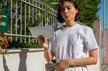 Fototapeta premium Surprised young woman in a white t-shirt holding an envelope near a mailbox on the street. A moment of curiosity or unexpected news, capturing emotion and everyday life in an outdoor setting.