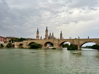 Obraz premium Puente de Piedra spans the Ebro River in Zaragoza, Spain. The stone bridge features multiple arches and leads to the Basilica de Nuestra Señora del Pilar in the background. 